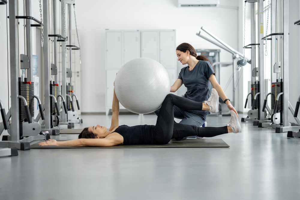 Woman doing exercises with fitness ball with rehabilitation specialist at the gym. Concept of physical therapy for health and recovery. Idea of recovery after pregnancy