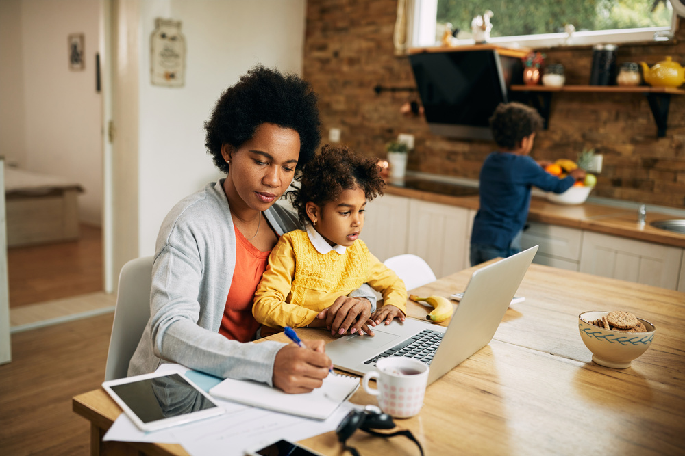 Black working mother taking notes while daughter is sitting on her lap and using laptop at home. Small boy is in the background.