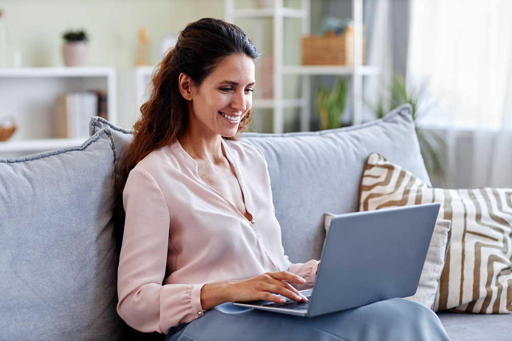 Portrait of smiling young woman using laptop while sitting on sofa at home and using internet or working online