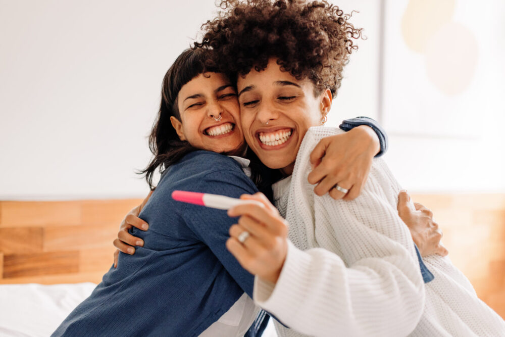 Celebrating having a baby on the way. Excited young lesbian couple smiling cheerfully and embracing each other after taking a home pregnancy test. Young female couple expecting a baby.