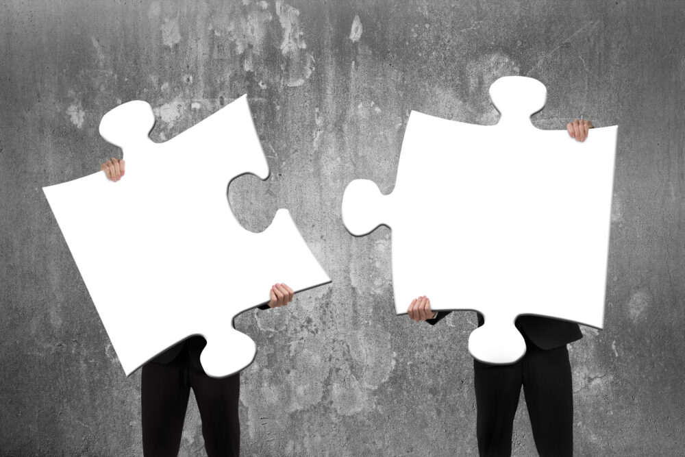 Two business people assembling blank white jigsaw puzzles with concrete wall background
