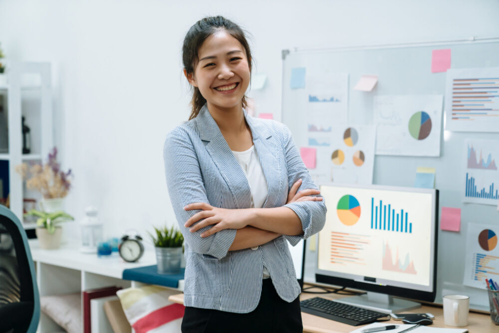 Young asian chinese woman freelancer stay indoors home office concept. confident female worker in smart casual wear with arms crossed face camera smiling with white board and documents on background.