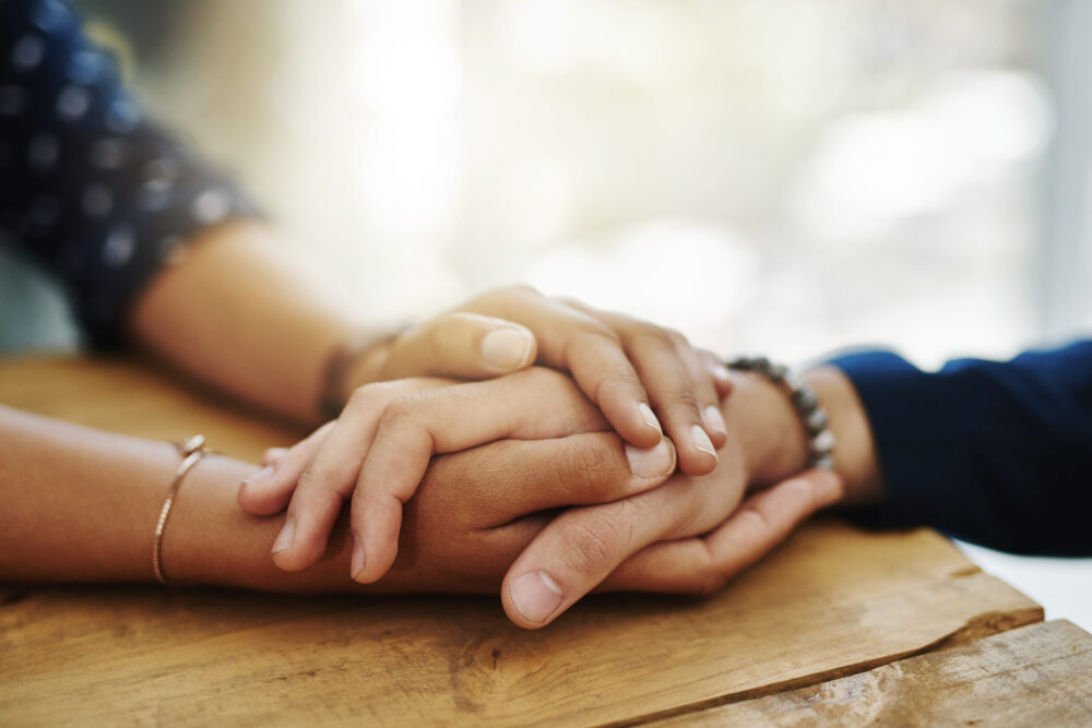 Holding hands, support and closeup with trust, solidarity and community on a home table. Therapy, diversity and gratitude of friends together with hope, respect and love for grief empathy and forgive.