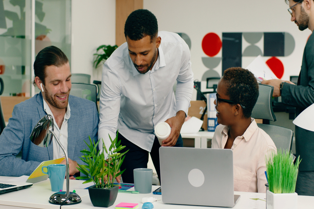 A very good looking black man is talking to his two co workers and showing some stuff on a paper on the desk, they are in a very nicely decorated office.