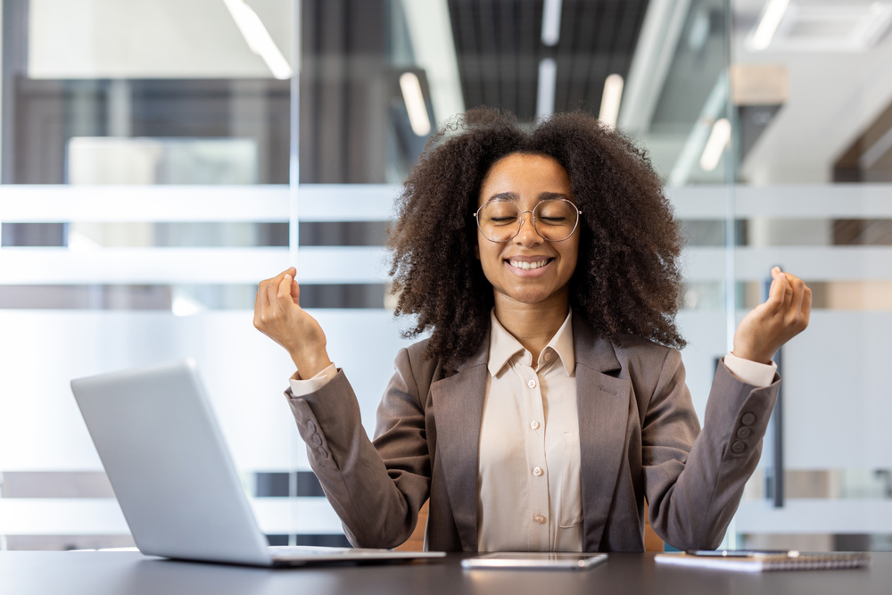 Close-up photo of a smiling young African-American woman in a suit sitting in an office at her workplace in the lotus position, meditating with her eyes closed