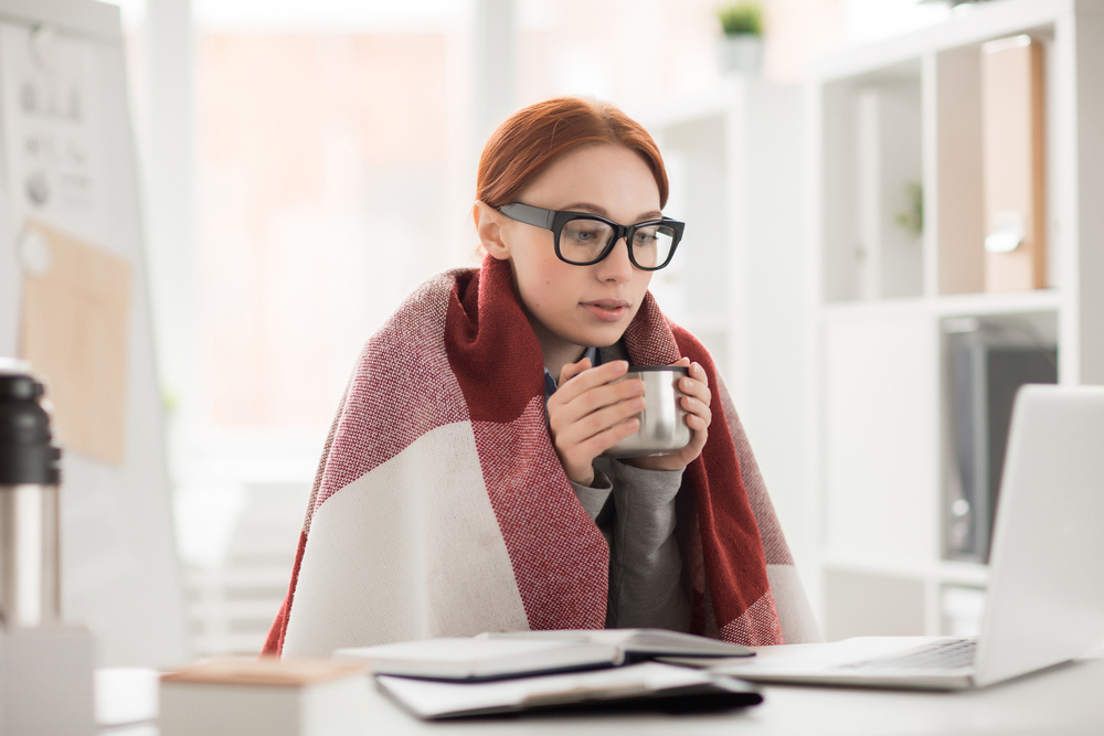 Young woman in plaid and eyeglasses drinking hot tea from mug during work in office