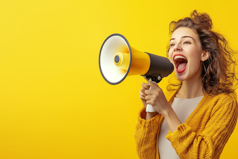 Happy young woman in a yellow sweater, announcing sale offers through a megaphone, isolated on a yellow background with copy space. Screaming into the megaphone. Studio portrait.