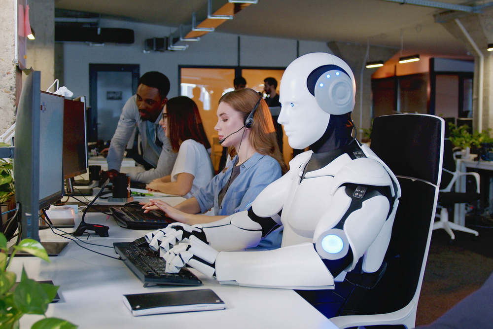 Robot working at computer among people. Maschine typing on keyboard in office. IT team of future. Futuristic worker. Humanoid work at call center. Support job. Selling concept. Technologies.