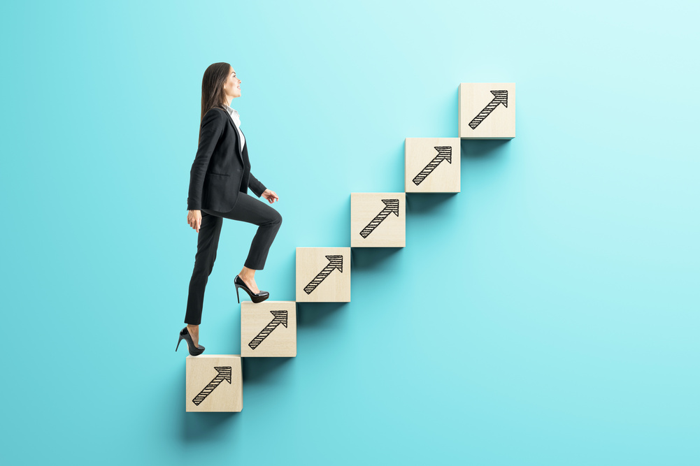 Confident businesswoman in formal attire climbing wooden blocks with arrows, symbolizing career growth, on a blue background. Concept of success