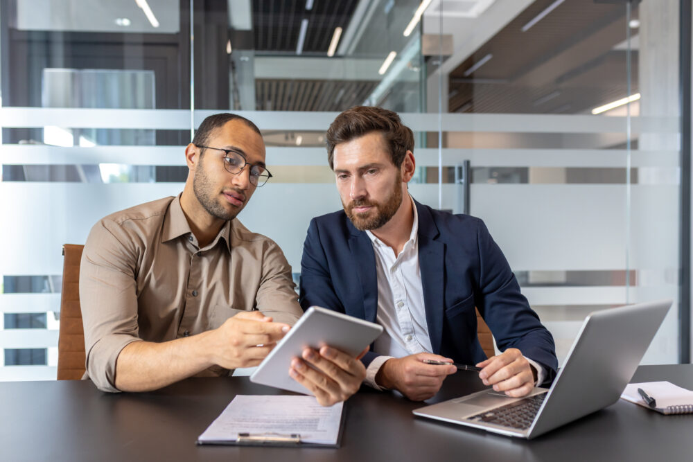 Two businessmen reviewing a tablet and laptop, discussing business strategies in a modern office setting.