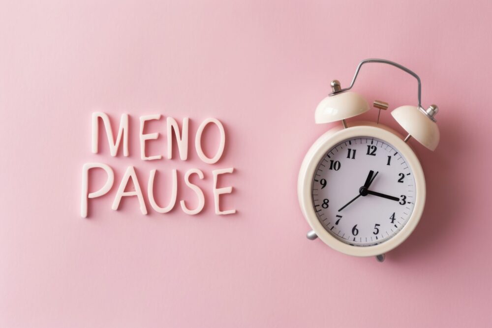 A symbolic representation of menopause featuring a white alarm clock on a pink backdrop, with the word 'MENOPAUSE' divided by a pause symbol