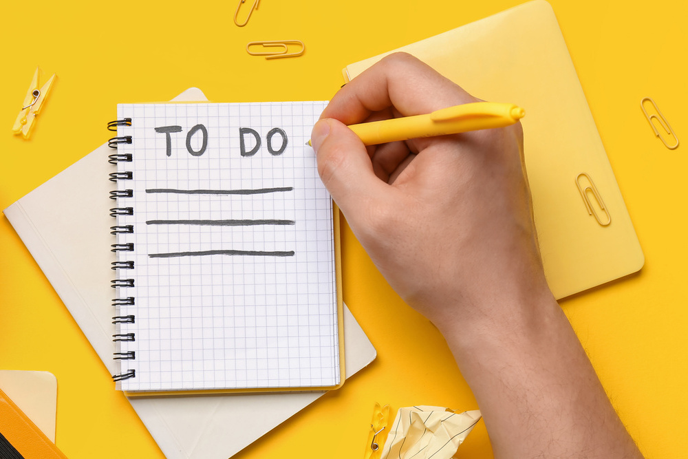 Hand with empty to-do list, notebooks, cup of coffee and office stationery on yellow background, top view
