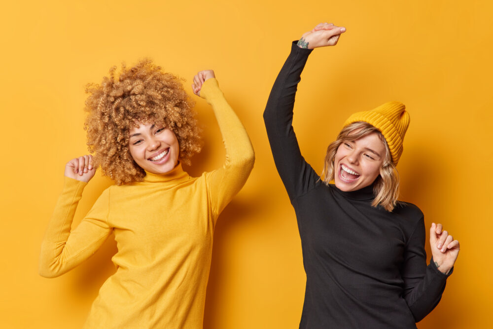 Positive cheerful young women have fun and dance carefree shake arms dressed in casual turtlenecks have glad expressions isolated over yellow background. People happiness and emotions concept
