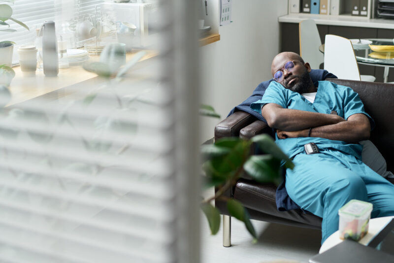 A healthcare worker in blue scrubs sleeps on a couch in a break room, conveying exhaustion. The room has plants and is partially visible through blinds.