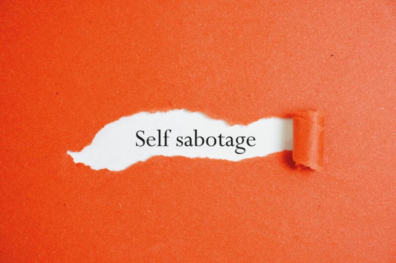 A torn sheet of paper revealing the words “self-sabotage” underneath, symbolising hidden behaviours that undermine performance and wellbeing