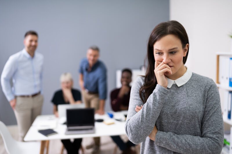 Employee looking concerned in office while colleagues display intimidating behaviour, representing workplace bullying and toxic culture