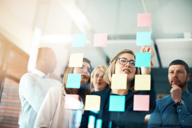 A team of professionals brainstorming workplace wellbeing initiatives using sticky notes on a glass wall during a collaborative planning session.