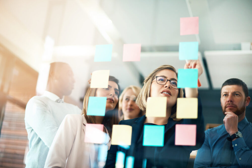 A team of professionals brainstorming workplace wellbeing initiatives using sticky notes on a glass wall during a collaborative planning session.