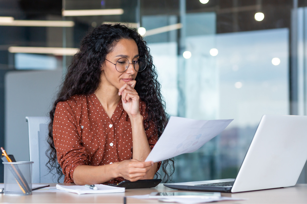 A woman in glasses, wearing a polka-dotted brown blouse, sits thoughtfully at a desk, examining a paper beside a laptop