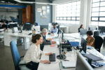 Employees working at desks in a busy open-plan office, illustrating the sedentary nature of modern work and the importance of building movement into the working day.
