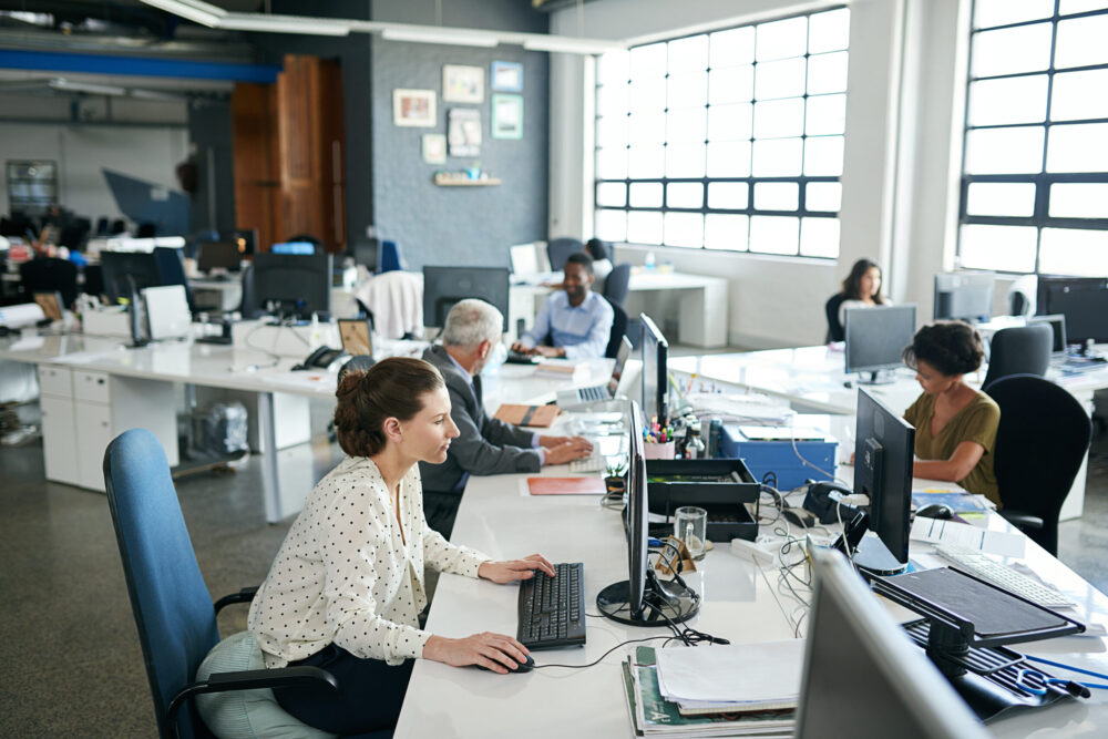 Employees working at desks in a busy open-plan office, illustrating the sedentary nature of modern work and the importance of building movement into the working day.