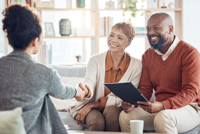A financial adviser shakes hands with a smiling couple seated in a bright living room, with a clipboard in hand.