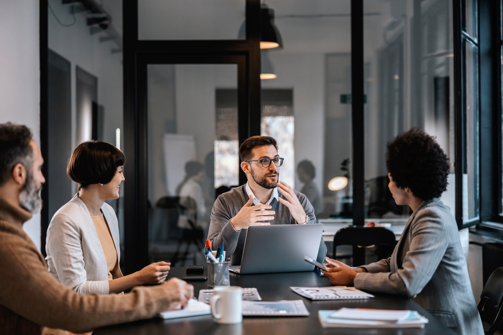 A group of professionals in a meeting room, discussing and collaborating around a table with a laptop and documents.