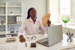 Young african american business woman employee using hand fan suffering from heat and summer high temperature working at the desk in office at her workplace.