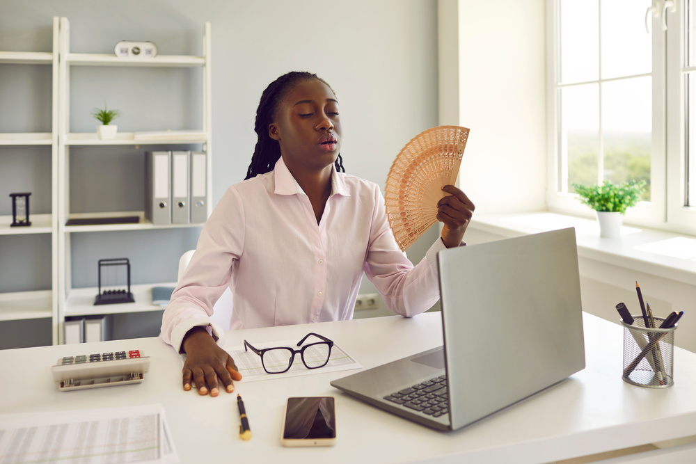 Young african american business woman employee using hand fan suffering from heat and summer high temperature working at the desk in office at her workplace.