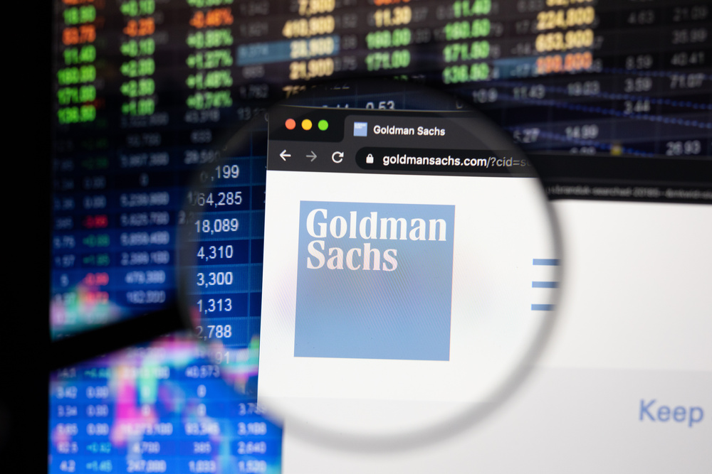 Goldman Sachs logo under a magnifying glass against a digital screen displaying fluctuating financial graphs.