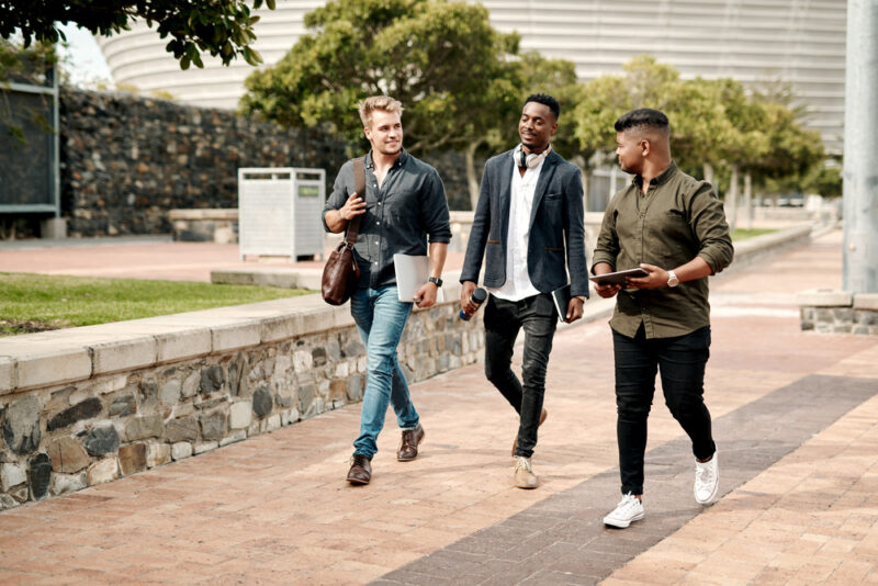 Three colleagues walking together outdoors carrying laptops and a tablet