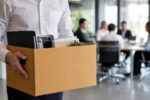 Employee holding a packed box of personal belongings in the foreground while colleagues take part in a team meeting in the background, illustrating organisational change and redundancy.