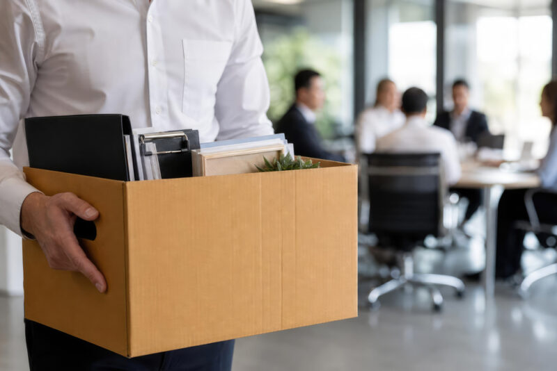 Employee holding a packed box of personal belongings in the foreground while colleagues take part in a team meeting in the background, illustrating organisational change and redundancy.
