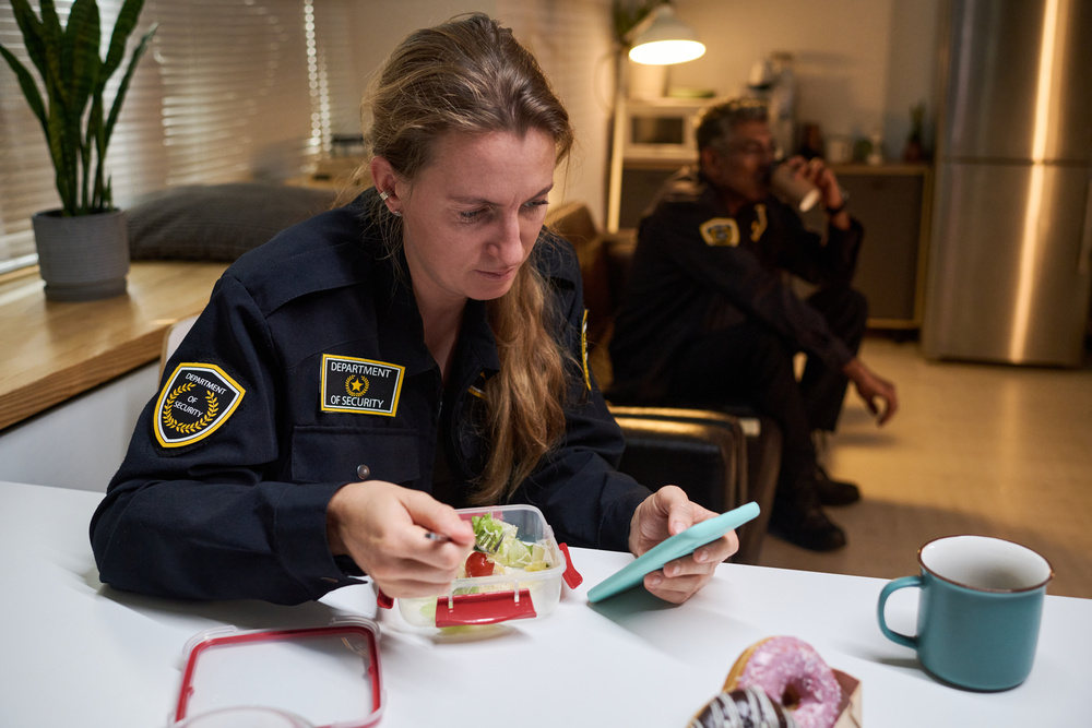 Caucasian security woman checking smartphone while eating meal during work break, colleague resting behind.