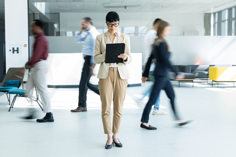 Businesswoman looking at a tablet in an office as colleagues walk past in the background