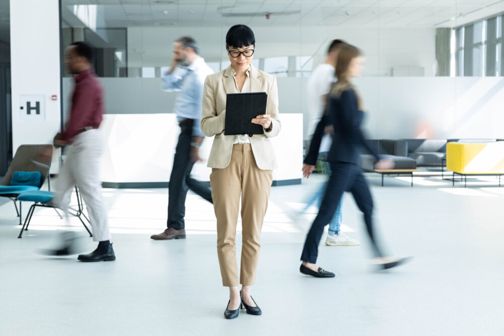 Businesswoman looking at a tablet in an office as colleagues walk past in the background